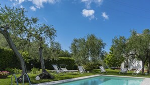 A rectangular swimming pool with clear blue water, surrounded by a grassy area and trees. There are several white lounge chairs and a small poolside table. - Alberto B by Interhome (Diano Marina)