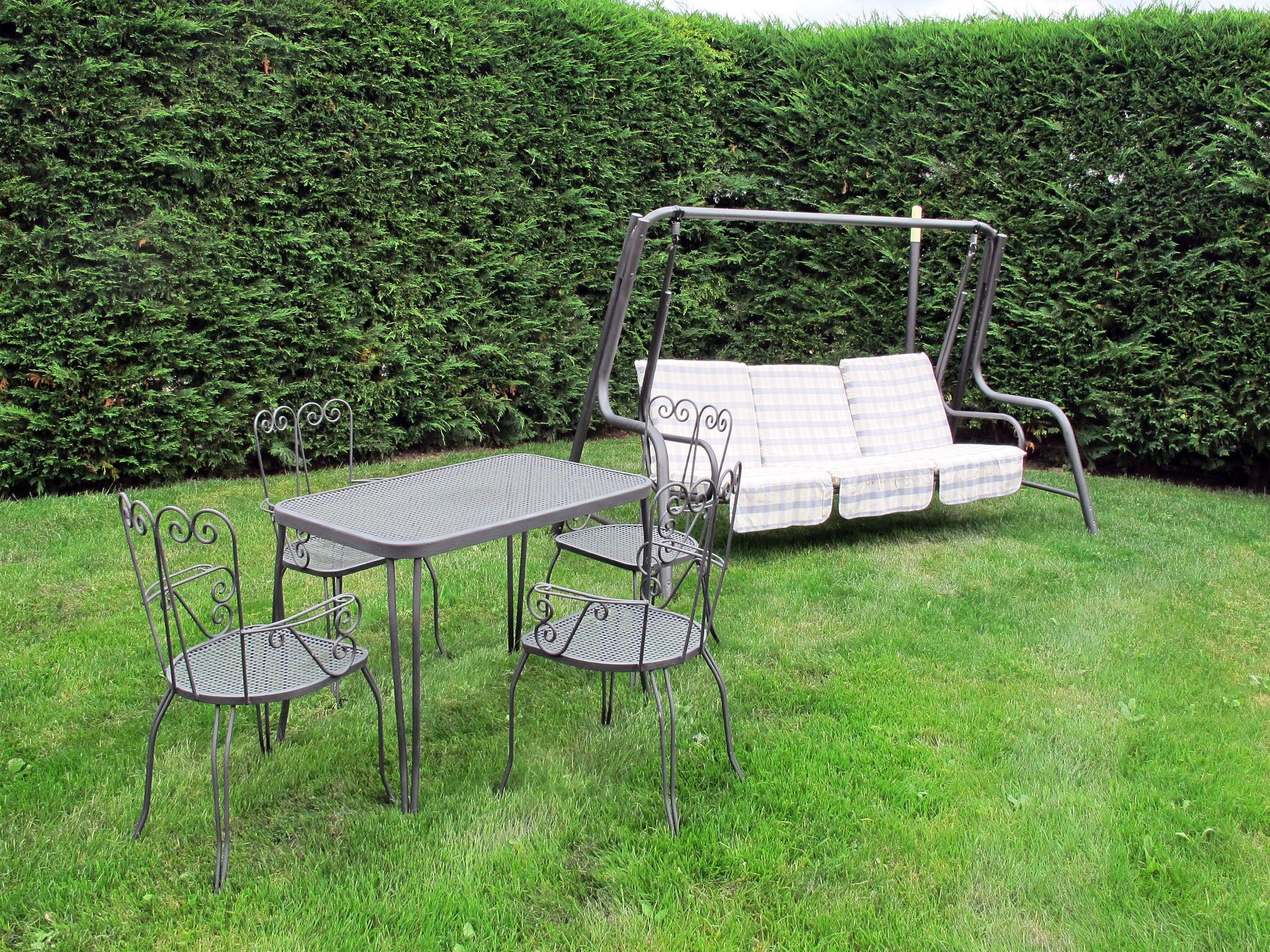 Een terras met een metalen tafel en stoelen, een schommelzit en een haag op de achtergrond.