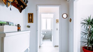 Un pasillo con pared blanca, piso de madera, una planta en maceta roja y una puerta que da a otra habitación.
