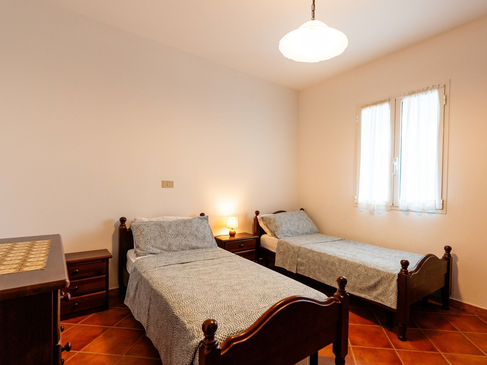A room with two wooden beds, a small bedside table with a lamp, a dresser, and a window with curtains.