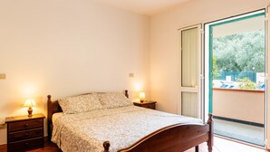 A bedroom with a wooden bed, two bedside tables with lamps, a sliding glass door to a balcony, and a view of greenery outside. - La Colombera by Interhome (Diano Marina)