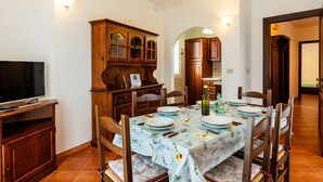 Uma sala de jantar com uma mesa posta para quatro pessoas, cadeiras de madeira, uma televisão e um armário com portas de vidro.