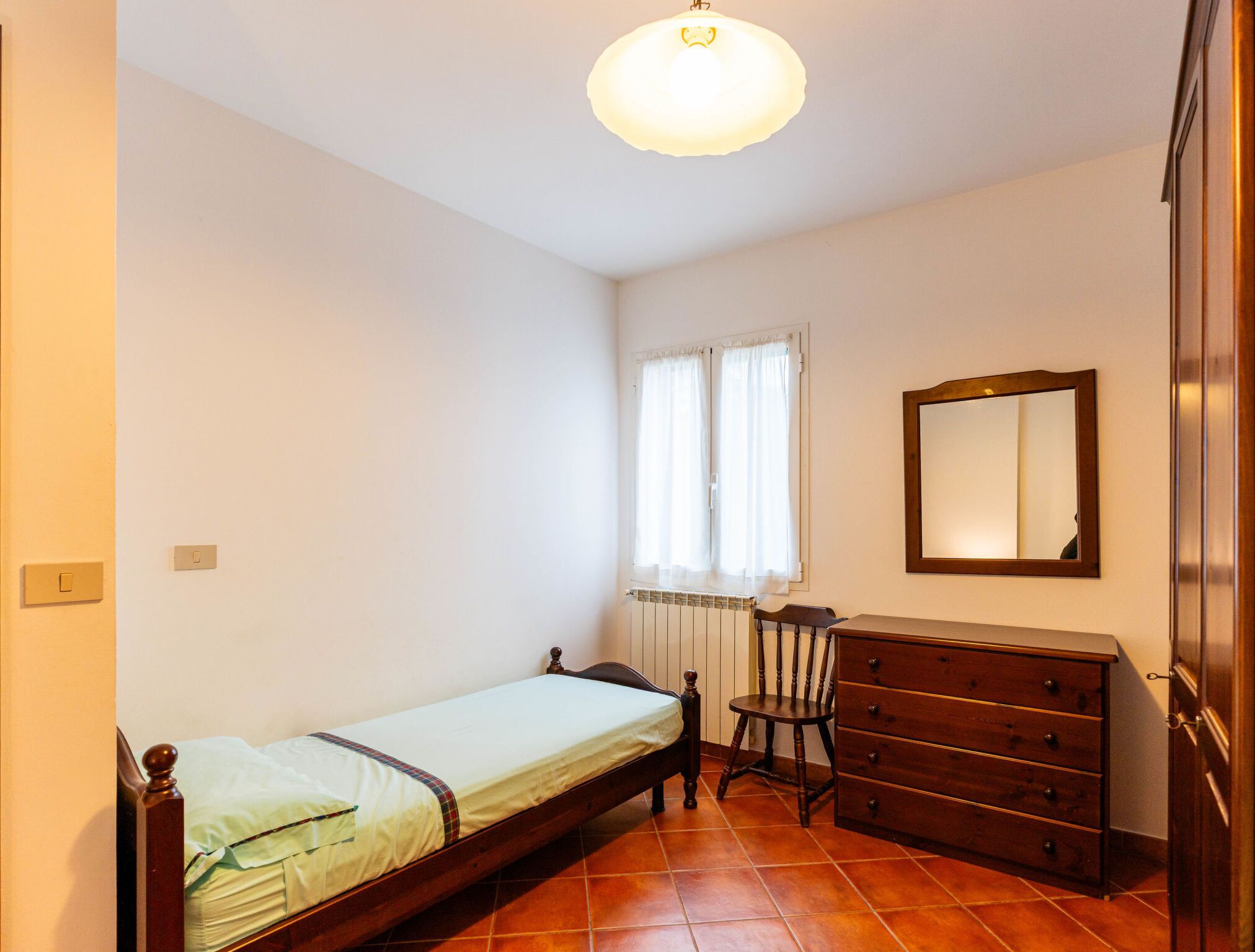 Une chambre avec un lit simple, une commode en bois, une chaise, un radiateur et une fenêtre avec des rideaux.