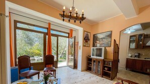 Amplia sala de estar con un gran ventanal que da a un jardín, comedor con una mesa dispuesta para dos, televisor sobre un soporte de madera y una cocina visible a través de un arco.