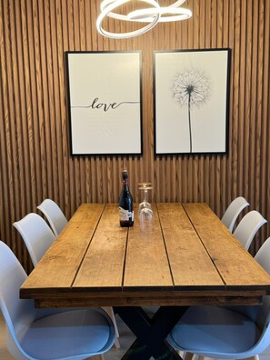 A wooden dining table with a bottle and glass, surrounded by white chairs, against a wooden wall with two framed pictures.