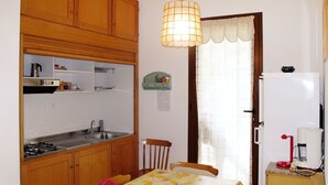 A compact kitchen with wooden cabinets, a gas stove, a sink, and a dining table with a floral tablecloth. - Ivano (PGI125) by Interhome (Poggi)