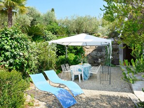 A covered outdoor seating area with lounge chairs and a table, surrounded by lush greenery and a stone wall.