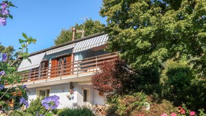 Una casa moderna de dos pisos con balcón, rodeada de vegetación exuberante y plantas florecientes.