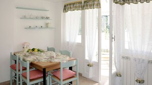 Ein Essbereich mit Tisch für vier Personen, hellblauen Stühlen und einer Tischdecke mit Blumenmuster. Über dem Tisch befinden sich Regale mit Küchenutensilien.