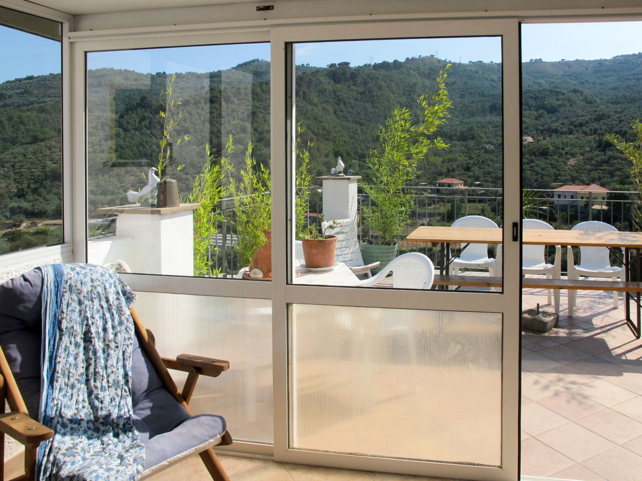 Un balcone con vista su un paesaggio montuoso, un tavolo con sedie e una poltrona con una coperta.