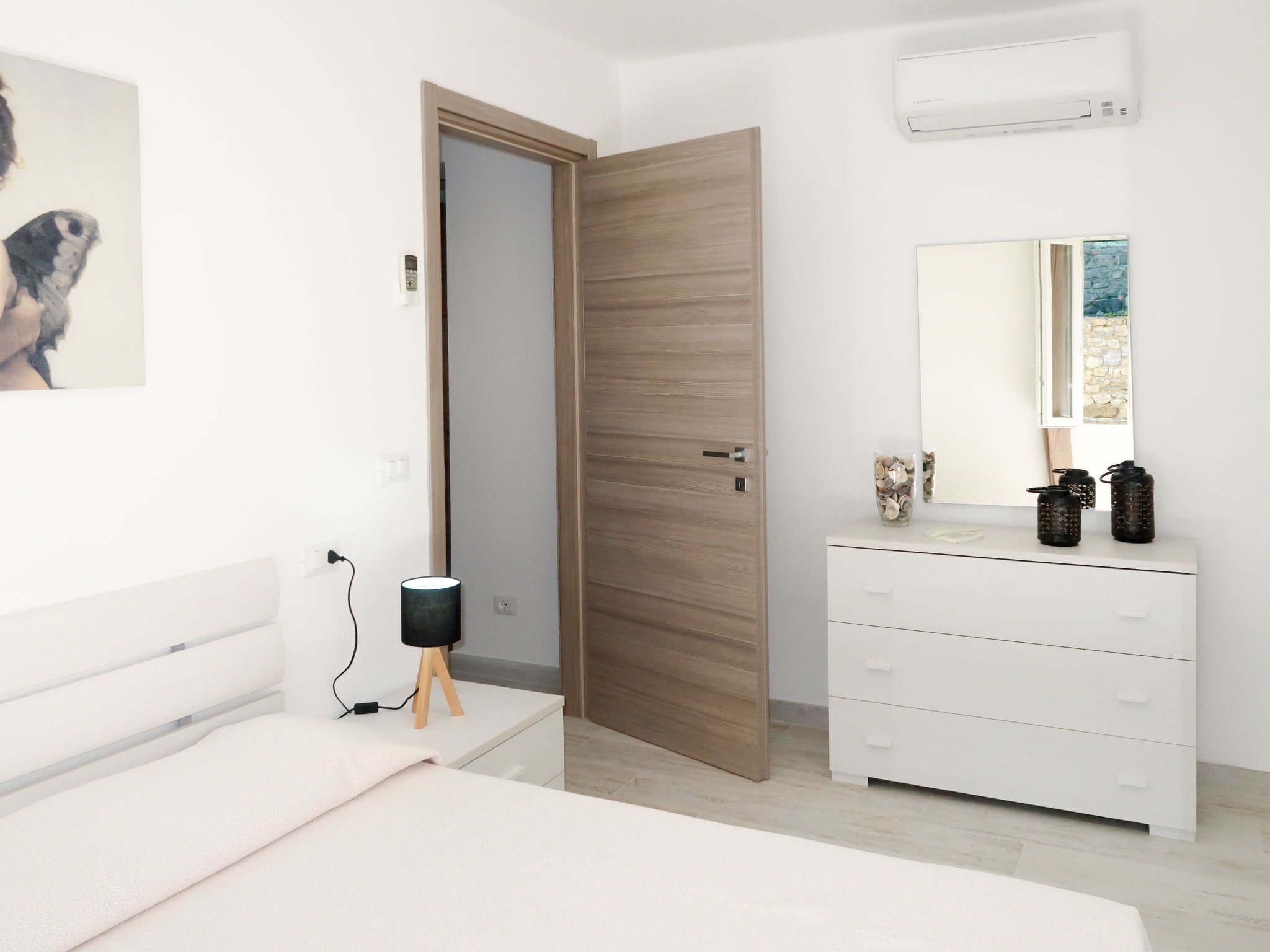 A modern bedroom with a wooden door, a white dresser, a bedside table, and a wall-mounted air conditioner.