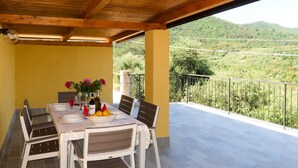 Uma área de refeições ao ar livre coberta, com uma mesa posta para dois, cadeiras e vista para uma paisagem montanhosa.