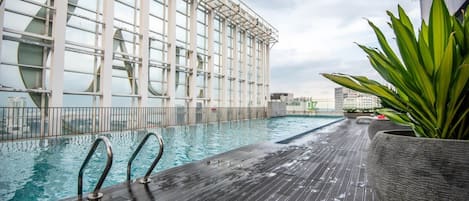 Piscina en la azotea con un deck de madera, rodeada por un edificio con fachada de vidrio.