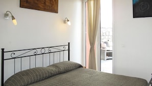 A bed with a metal frame and a grey quilt, a wall-mounted light fixture, and a view of the outside through a door. - Ciliegi (SLR354) by Interhome (San Lorenzo al Mare)