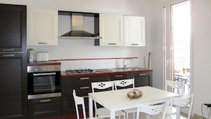 A compact kitchen with a gas stove, oven, and white cabinets. There is a dining table with four chairs and a window providing natural light. - Ciliegi (SLR354) by Interhome (San Lorenzo al Mare)