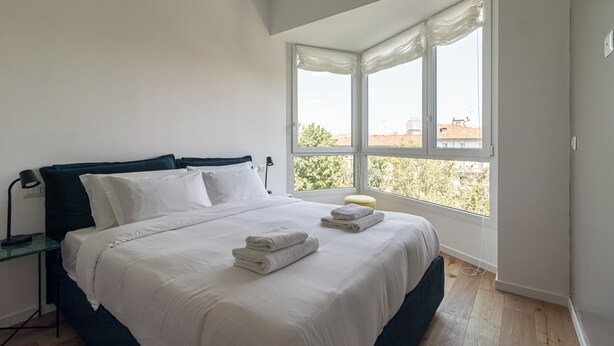 A bedroom with a bed, a nightstand, a lamp, and a window with a view of buildings and trees. - Italianway Comfort - D'aragona 2 (Milan)