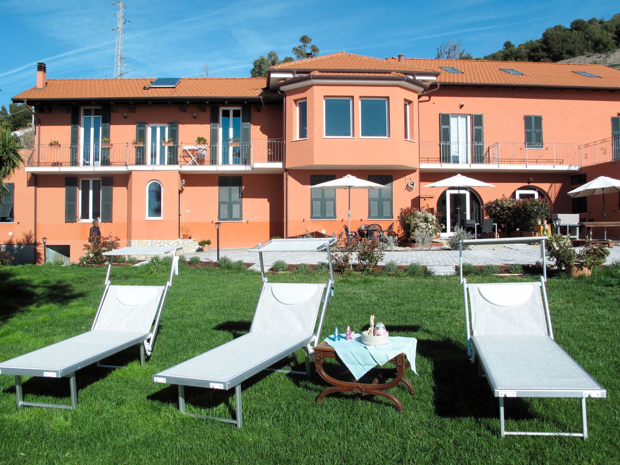 Un bâtiment à deux étages, doté d’un toit recouvert de tuiles, d’un espace extérieur aménagé avec des chaises longues blanches et d’une petite table recouverte d’une nappe.