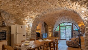 Uma cozinha rústica de pedra com uma mesa de jantar e cadeiras de madeira, uma geladeira e uma grande janela em arco.
