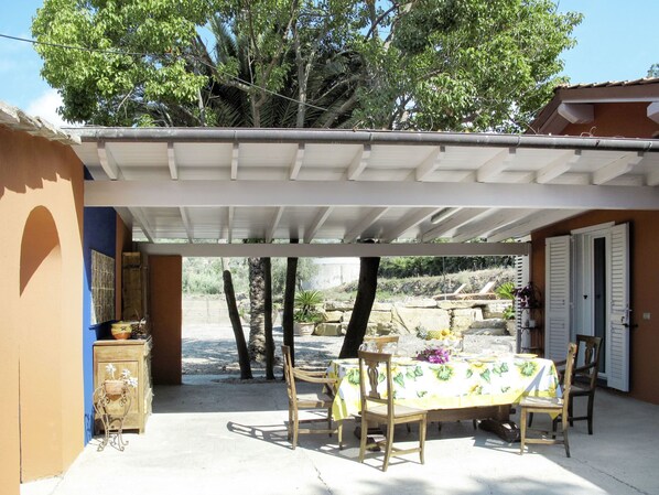 Un patio exterior con mesa y sillas cubiertas, rodeado de árboles y un muro de piedra.