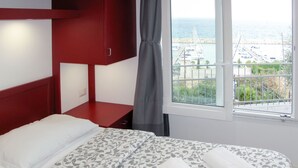 Una habitación de hotel con cama, un armario rojo, una ventana con vistas al mar y barcos, y una cortina.