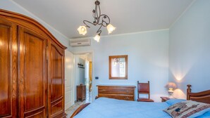 Ein Schlafzimmer mit einem großen Holzschrank, einem Bett mit blauer Steppdecke, einer Kommode, einem Stuhl, einem Nachttisch mit Lampe und einem Spiegel an der Wand.