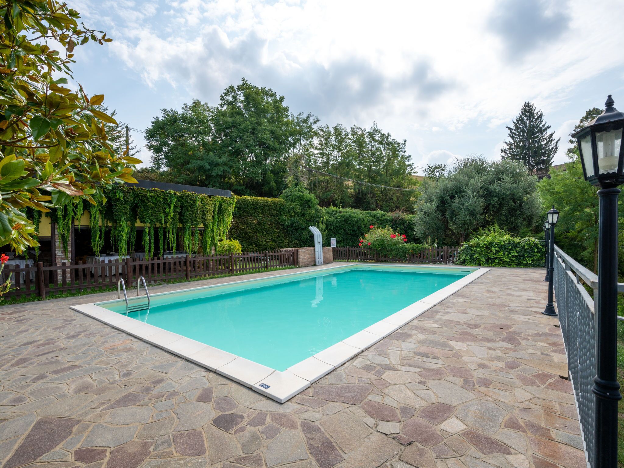 A rectangular swimming pool with stone tiles, surrounded by a wooden fence and greenery.