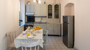 Une cuisine avec des meubles de couleur gris clair, un réfrigérateur en acier inoxydable, une table à manger recouverte d’une nappe en dentelle et une horloge murale.
