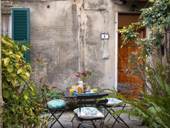 An outdoor seating area with a table set for two, surrounded by greenery and a rustic building facade. - Caruggiu (PTB193) by Interhome (Pietrabruna)