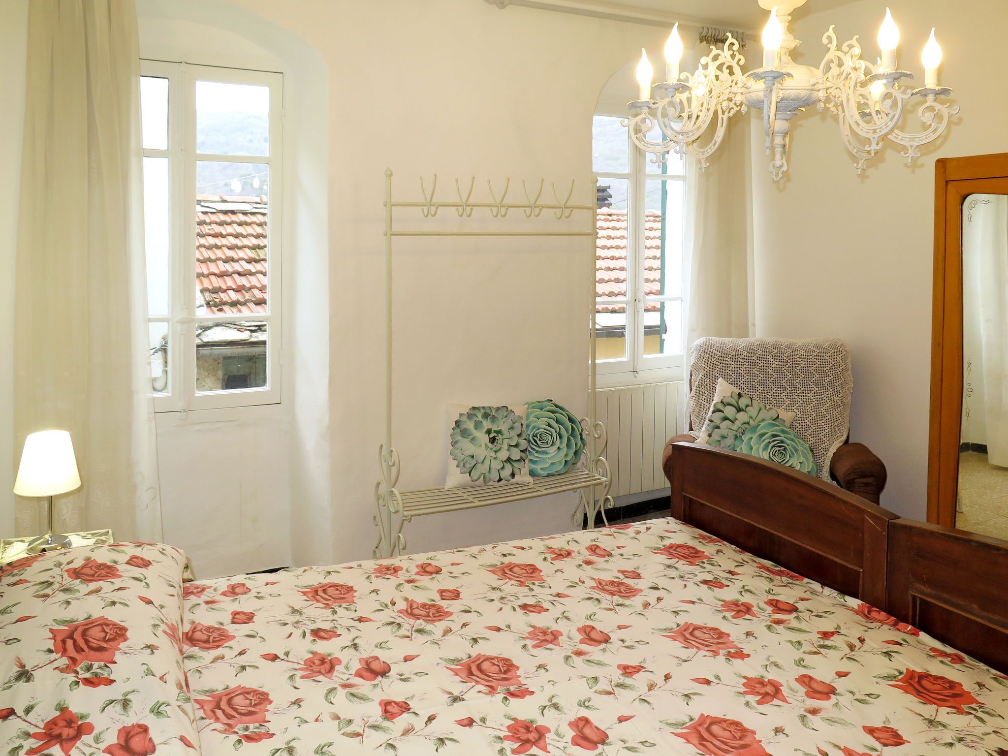 A bedroom with a floral bedspread, a white metal drying rack, a wooden chair with a lace cover, a chandelier, and a window with a view of a tiled roof.