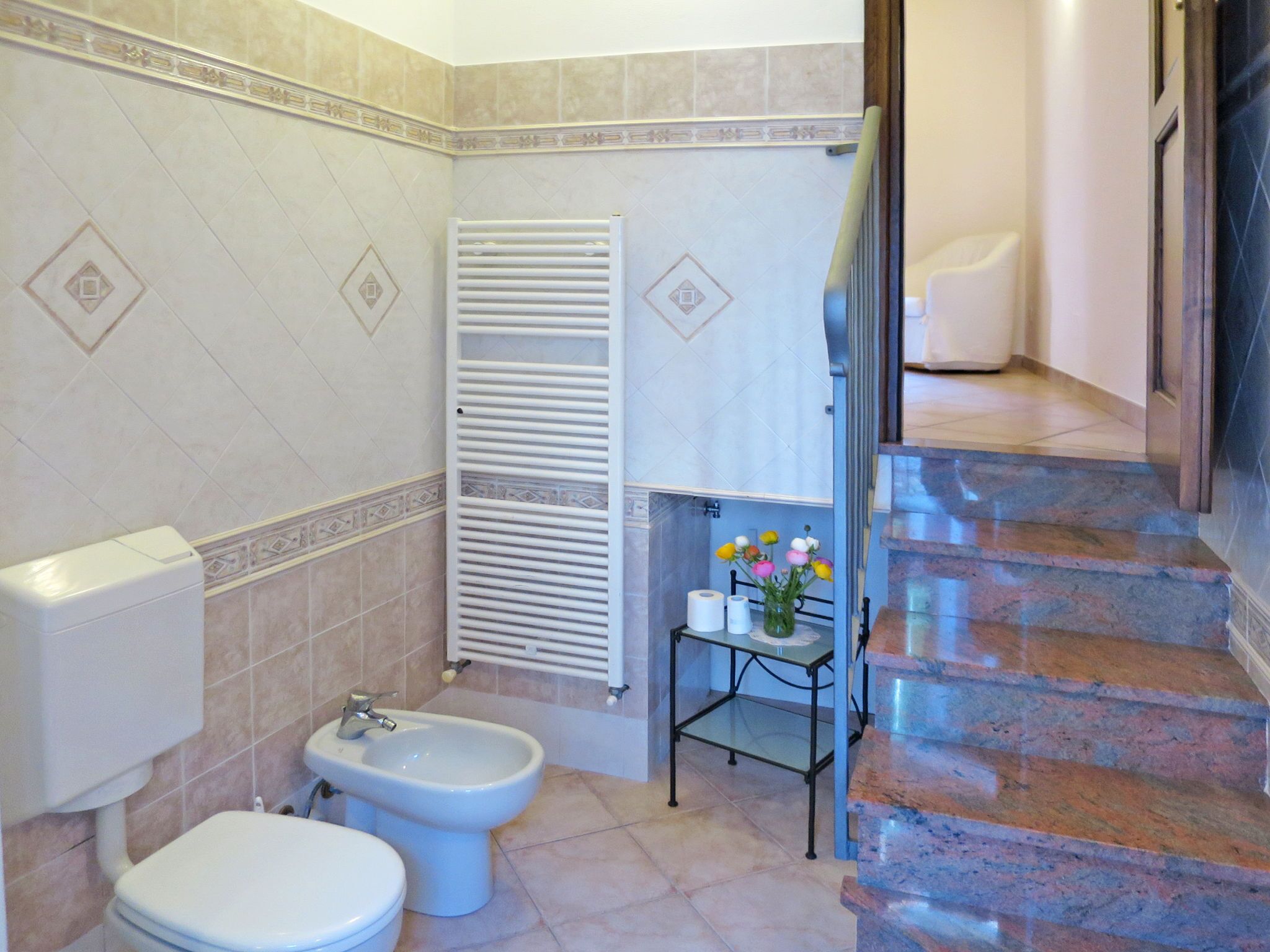 A bathroom with a toilet, bidet, towel rack, and a small table with a vase of flowers.
