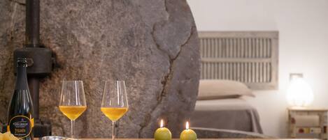 Uma parede de pedra, uma garrafa de vinho, duas taças de vinho, velas e uma mesa de madeira num quarto.