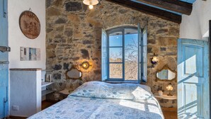 A rustic bedroom with a stone wall, a wooden ceiling, a bed with a patterned bedspread, a window with a view of trees, and a blue door. - Azzurra (PTB145) by Interhome (Pietrabruna)