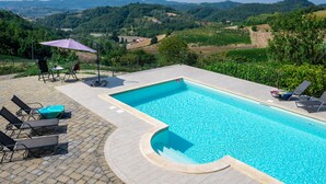 Una piscina rectangular con borde curvo, rodeada por una terraza pavimentada. Hay sillas de descanso y una mesa con sombrilla. La piscina tiene vistas a un paisaje montañoso con viñedos.