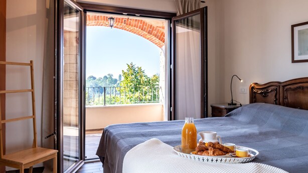Un dormitorio con una cama grande, una silla de madera, una mesita de noche con lámpara y un balcón con vistas a zonas verdes.