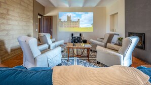 Una sala de estar moderna con una pantalla grande que muestra un castillo, asientos cómodos, chimenea y una pared de piedra.