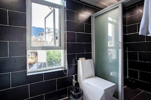 A bathroom with black tiles, a white toilet, a glass shower enclosure, and a window with a view of buildings and mountains. - Xiaochengshanyue Inn ((Dali Ancient City South Gate) (Dali City)