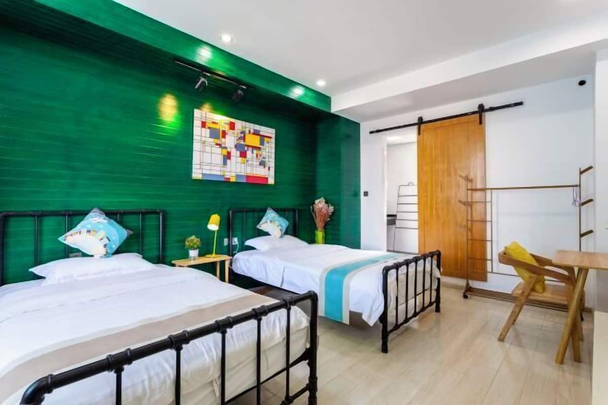 A room with two beds, a desk, a chair, a wooden door, and a green wall with a colorful artwork.