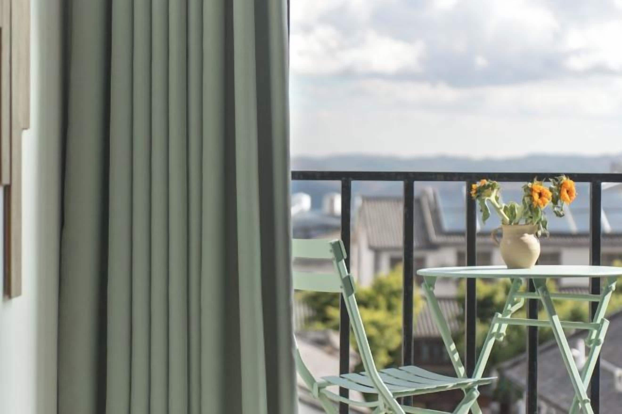 A balcony with a table and chairs, a vase with flowers, and a view of buildings and a cloudy sky.