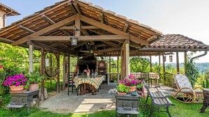 Un patio rústico al aire libre con una pérgola de madera, una mesa con mantel a cuadros, plantas en macetas y una estufa antigua.