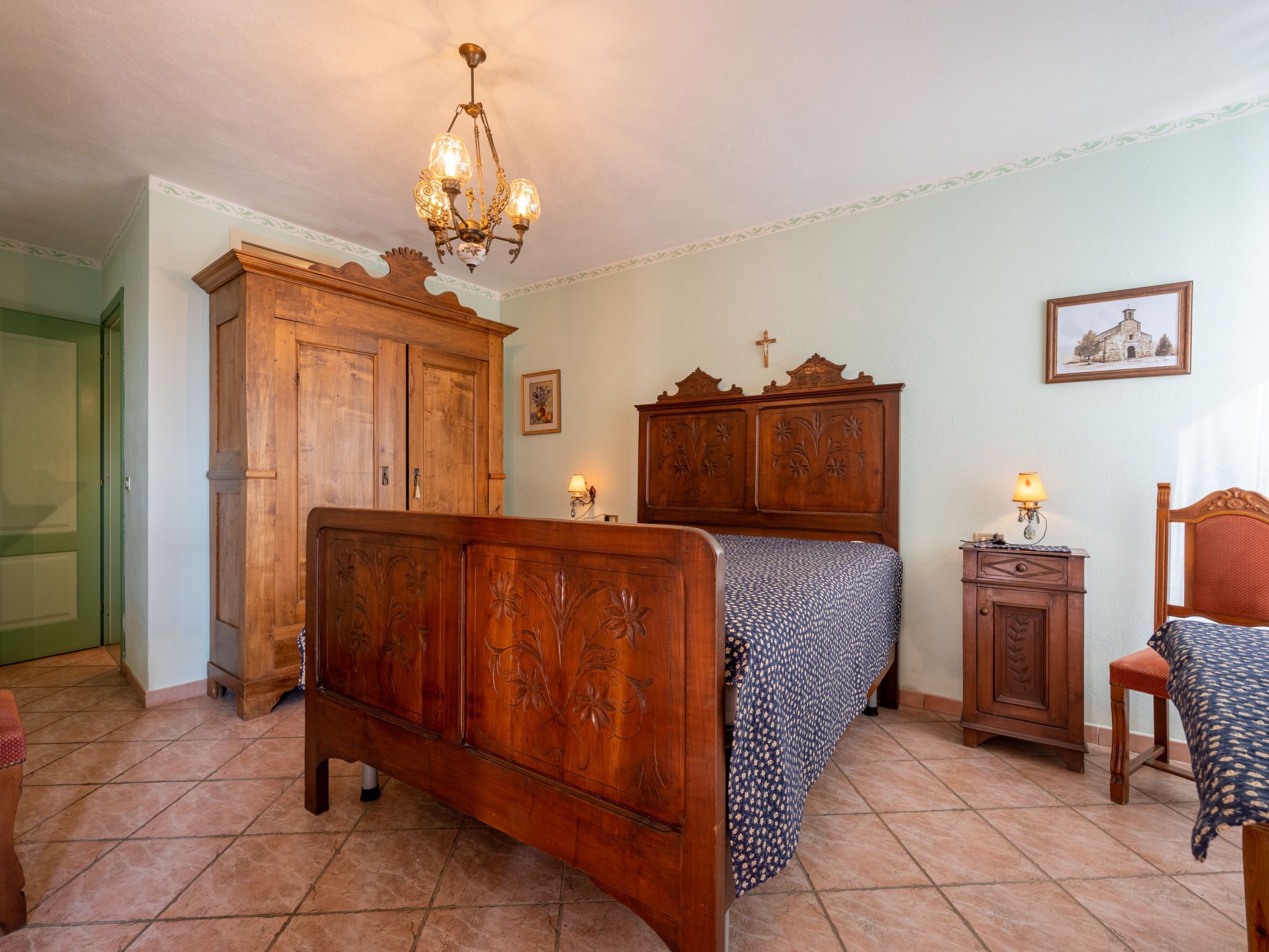 A bedroom with a wooden bed, a nightstand, a dresser, a chandelier, and a cross on the wall.