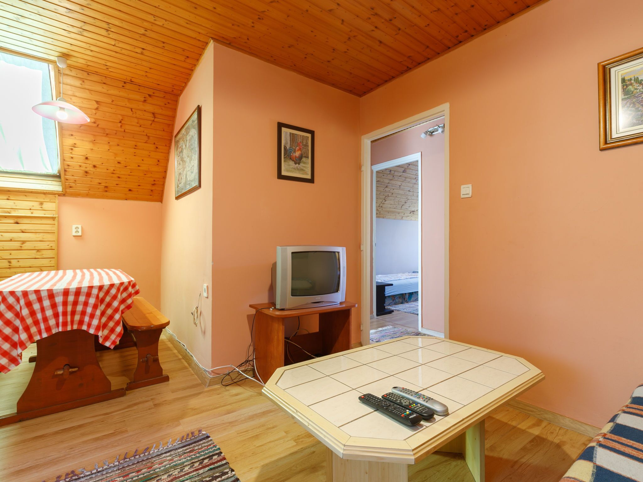 Ein gemütliches Zimmer mit Holzbalkendecke, einem Tisch mit kariertem Tuch, einem Fernseher auf einem Schrank und einem gefliesten Couchtisch.