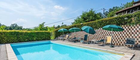 Une piscine rectangulaire bordée de carreaux, entourée de chaises longues et d’ombrelles vertes, le tout se dressant sur fond de haie et de mur de briques.