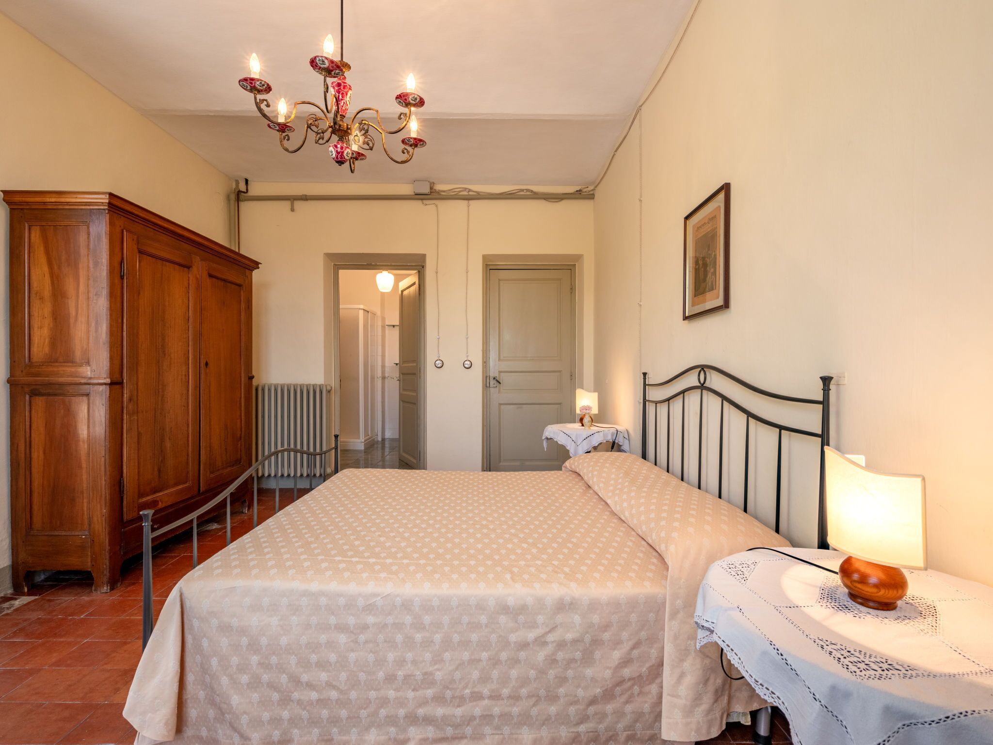 A bedroom with a large bed, a wooden wardrobe, a chandelier, and a small table with a lamp.