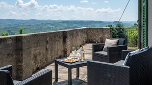 Un balcón con mobiliario de mimbre, una mesa con comida y bebidas, y una vista panorámica de colinas ondulantes.