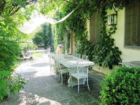 Une terrasse extérieure avec une table et des chaises blanches, entourée de verdure et agrémentée d’une pergola.