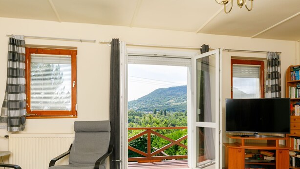 Un salón con un sofá gris, un televisor sobre un mueble de madera y vistas a un paisaje verde a través de una puerta corredera de cristal.