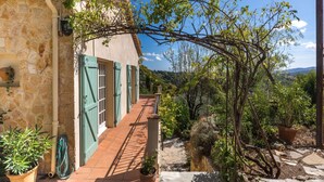 A stone building with a tiled pathway leading to a garden area with potted plants and a view of the countryside. - La Garance by Interhome (Grasse)