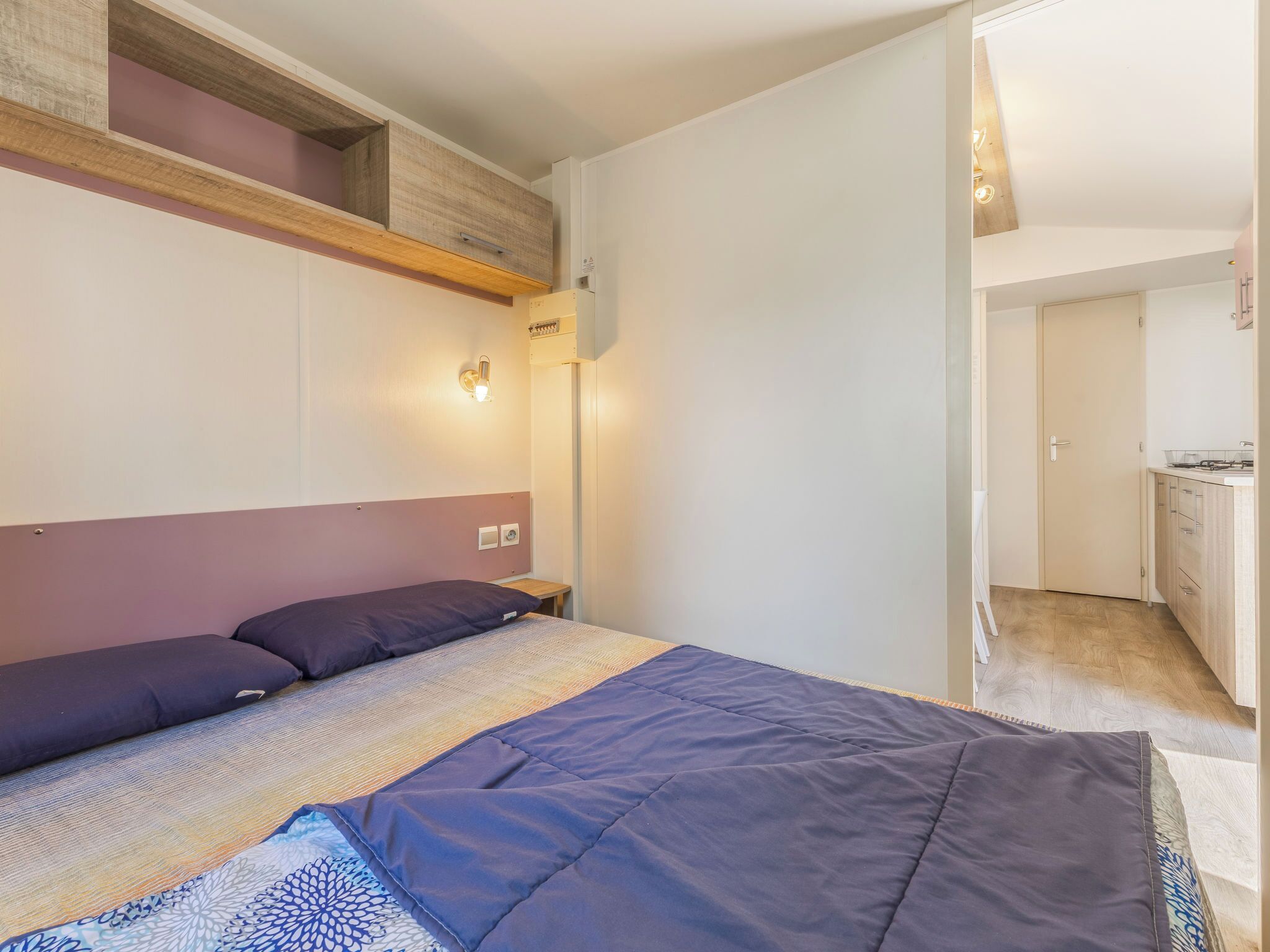 Ein Schlafzimmer mit Bett, Nachttisch, Wandleuchte und Blick zur Küche.