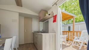 Une cuisine compacte dotée d’un plan de travail en bois, un coin repas avec des chaises blanches et une terrasse en bois avec des sièges d’extérieur.