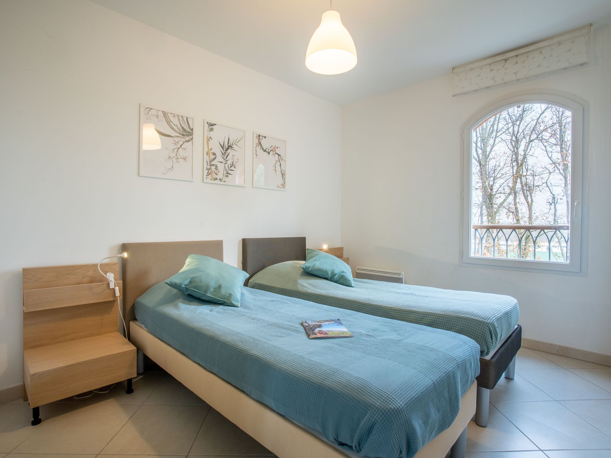 A bedroom with two beds, a nightstand, a bedside table, a window with a view, and wall art.